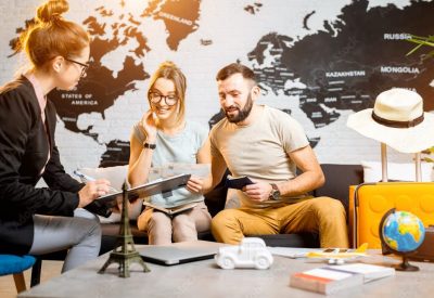 Travel planning session: two adults look at tickets with a consultant in a modern travel office, world map wall behind them.
