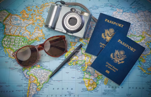 Camera, two blue passports, and sunglasses laid on a world map for travel planning or packing. Nylon strap visible on camera.