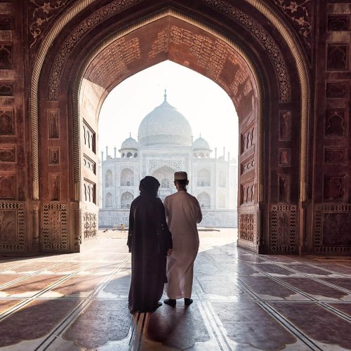 A young couple face away from the camera and pass under a large stone walkway to enter the stunning Taj Mahal in India.