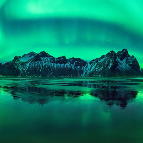 A tall and snowy mountain in Iceland surrounded by a beautiful ocean being cruised by Sky Bird Travel & Tours Sky Vacations. In the background, the beautiful natural phenomenon of the aurora borealis, or the northern lights, brightens the night sky with green hues.